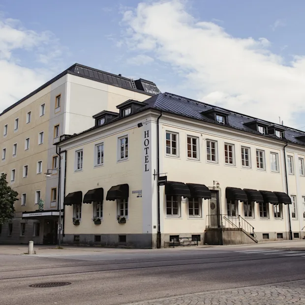 En kremfarget hotellbygning med svarte markiser på et gatehjørne under en delvis skyet himmel. Ordet HOTEL er synlig på siden.