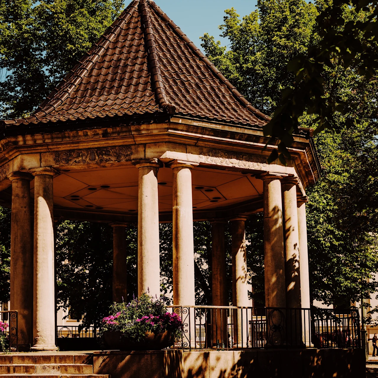 A gazebo with a tiled roof stands surrounded by trees, with sunlight casting shadows. Flowers adorn a railing in a peaceful park setting.