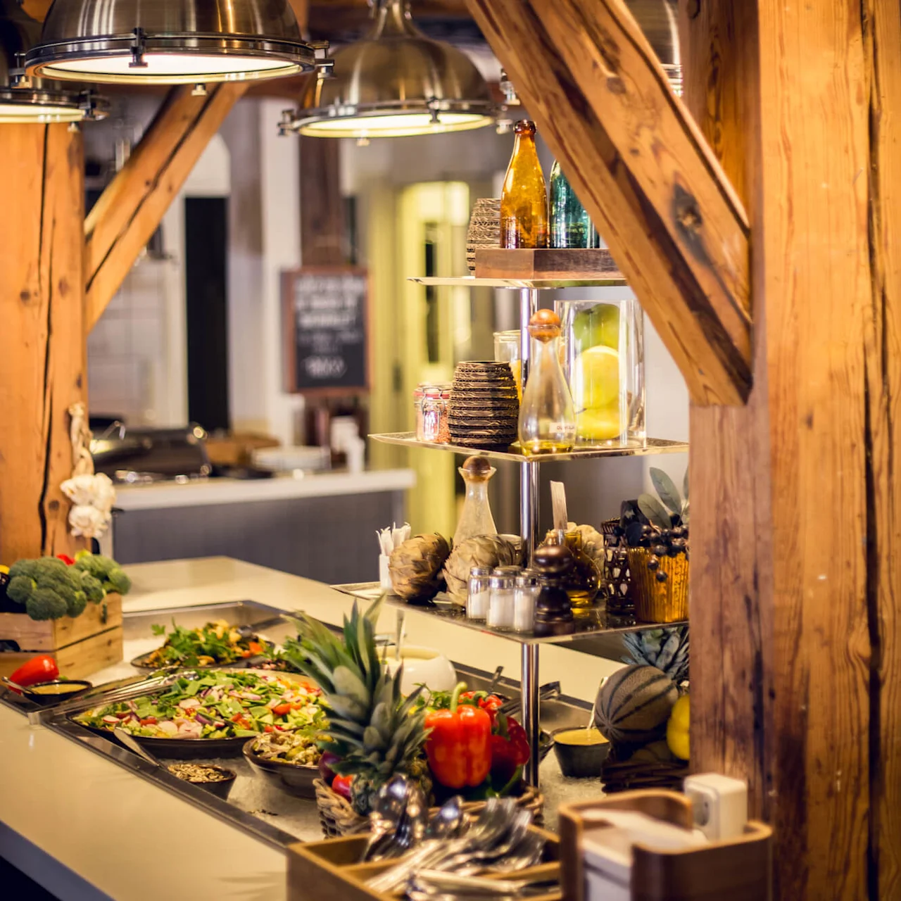 A buffet spread with salads, fruits, and cakes under metal lamps, framed by wooden beams inside a cozy dining area.