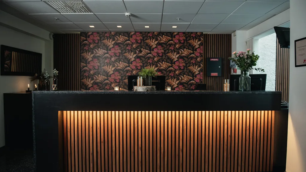 En receptionsdisk har träpaneler och varm belysning undertill. Bakom finns en vägg med blommönster och växter, med ett modernt tak och dekorativa element runtomkring.