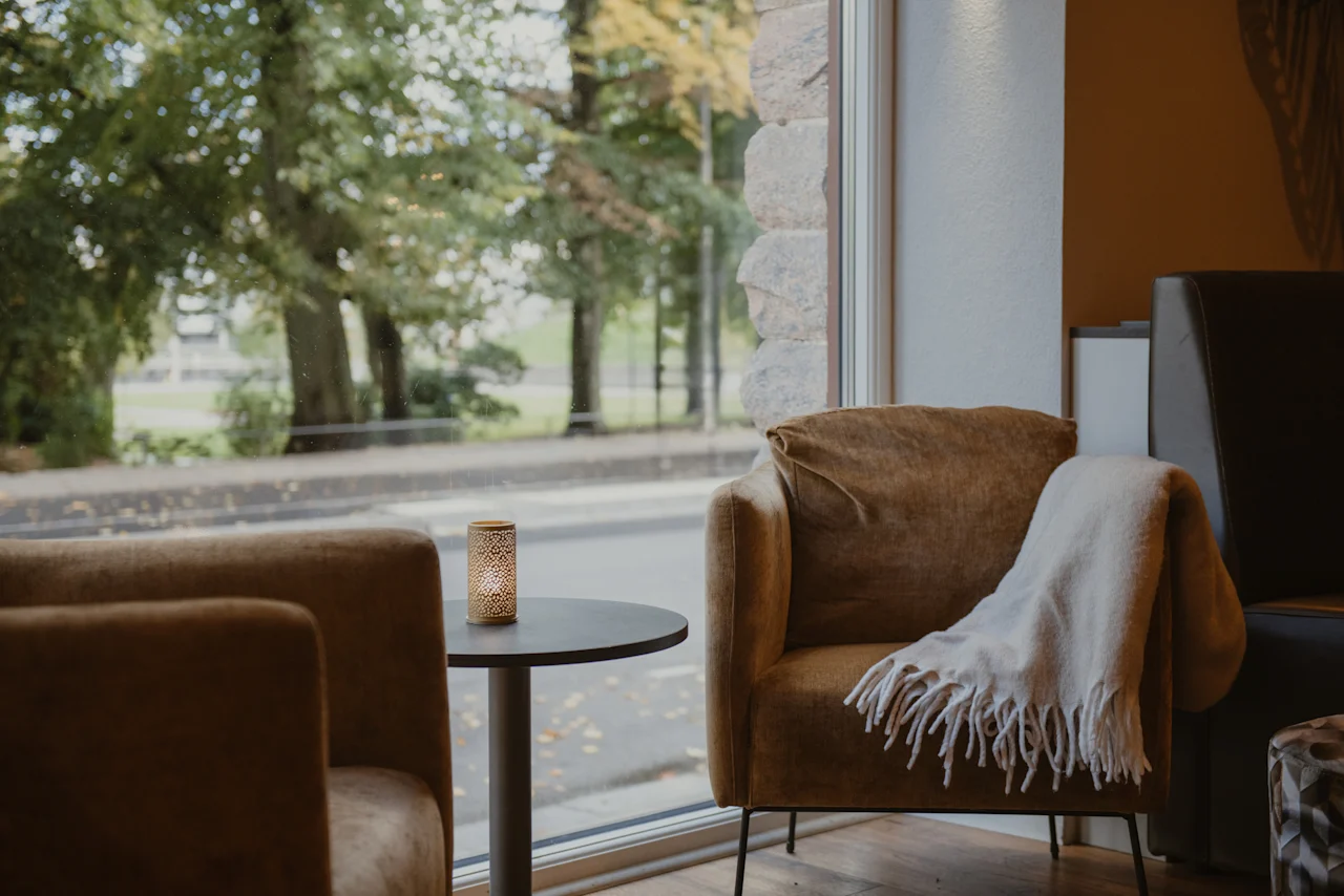 Et hyggeligt siddeområde med to lænestole, et lille bord med et tændt lys og et tæppe, ved siden af et vindue med udsigt over en træbeklædt gade.