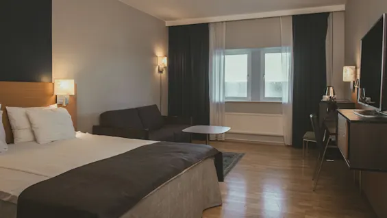 A bed with white sheets and dark blanket stands in a hotel room, alongside a sofa, table, window with curtains, and a TV on a wooden dresser.