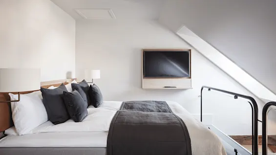 A neatly made bed with white and gray bedding sits in a minimalist attic room. A television is mounted on the sloped wall opposite, near a black railing.