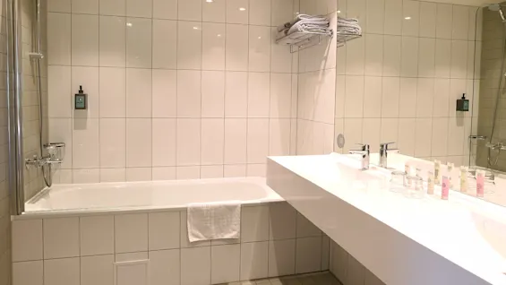 Et hvidt flisebelagt badeværelse har et badekar med en vægmonteret bruser, et stort spejl over en moderne håndvask med toiletartikler, og håndklæder stabler på en hylde.