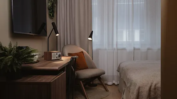 A cozy bedroom features a bed with gray linens, a wooden desk with a vintage radio and lamp, and a comfy chair with an orange cushion. A window provides natural light.