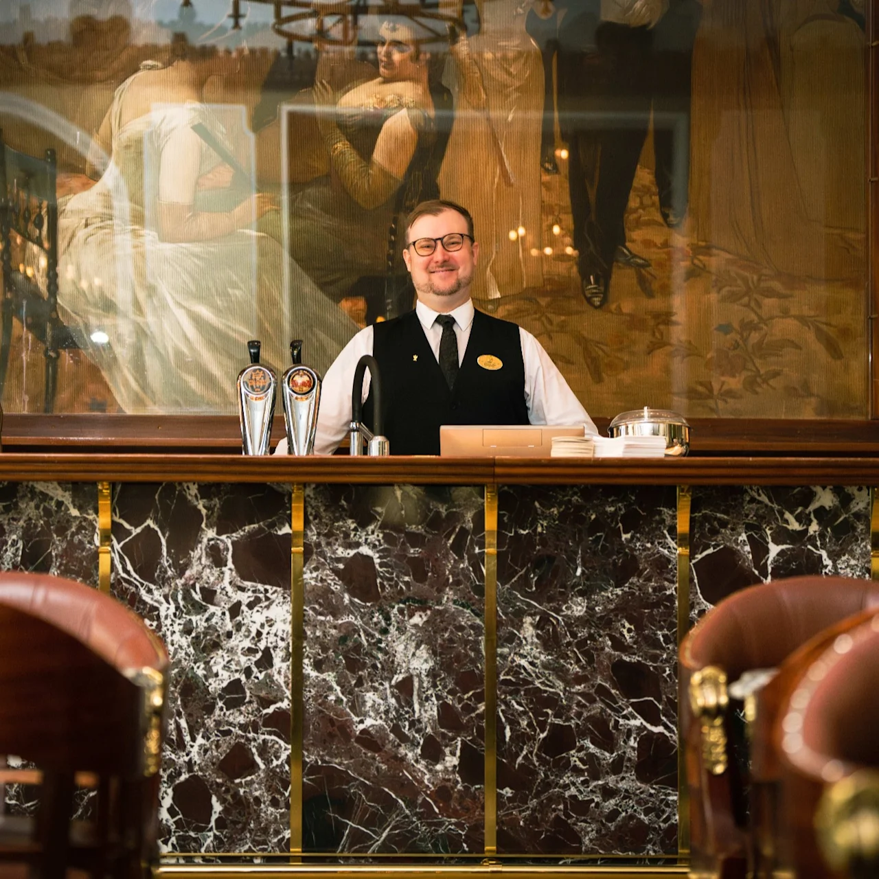 A smiling bartender stands behind a marble bar counter in a classic, elegant bar with paintings and wooden furnishings at Hotel Kämp in Helsinki.