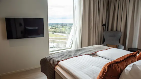 A bed with orange pillows and gray bedspread sits near a large window overlooking greenery. A wall-mounted TV and an armchair with a lamp are nearby.