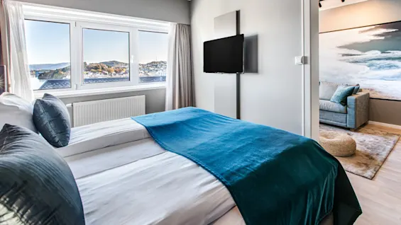 En pent seng med turkise sengeklær vender mot en veggmontert TV i et moderne soverom. Store vinduer viser en naturskjønn utsikt. Tilstøtende rom har sofa og havtema.