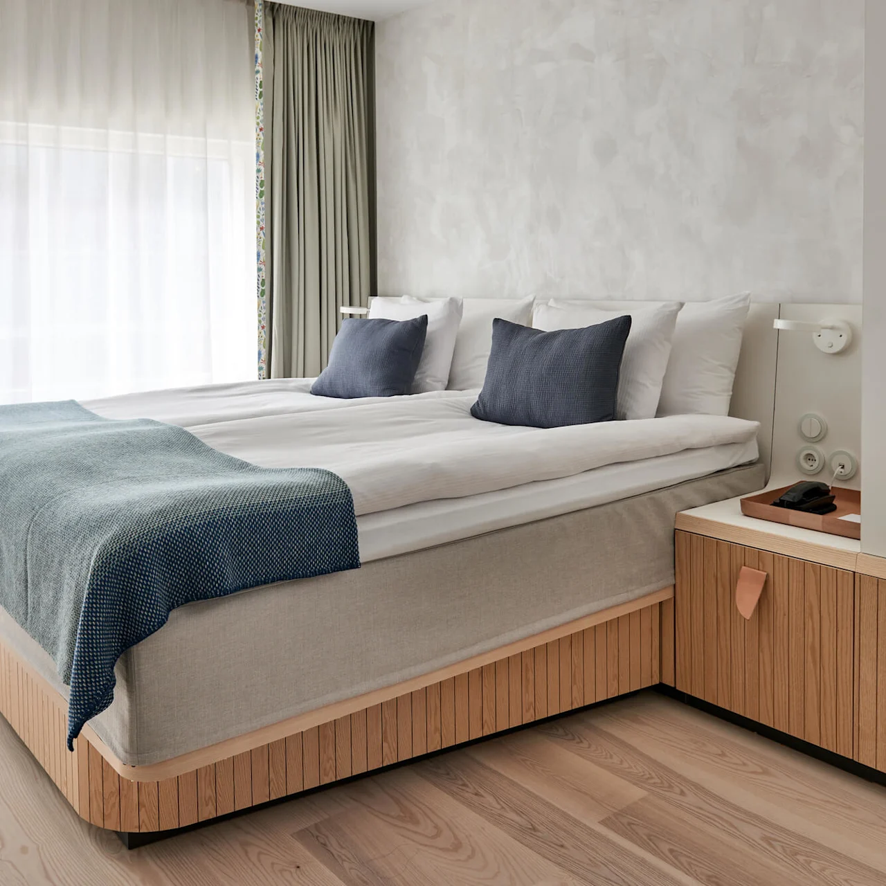 A neatly made bed with blue pillows and blanket rests in a bright, minimalist bedroom featuring light wood furniture and large, sheer-covered windows.