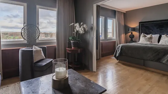 A cozy hotel room with a neatly made bed, soft pillows, and a nightstand with a lamp. A chair with decorative pillows sits by a window, draped with curtains. A small table holds an orchid plant.
