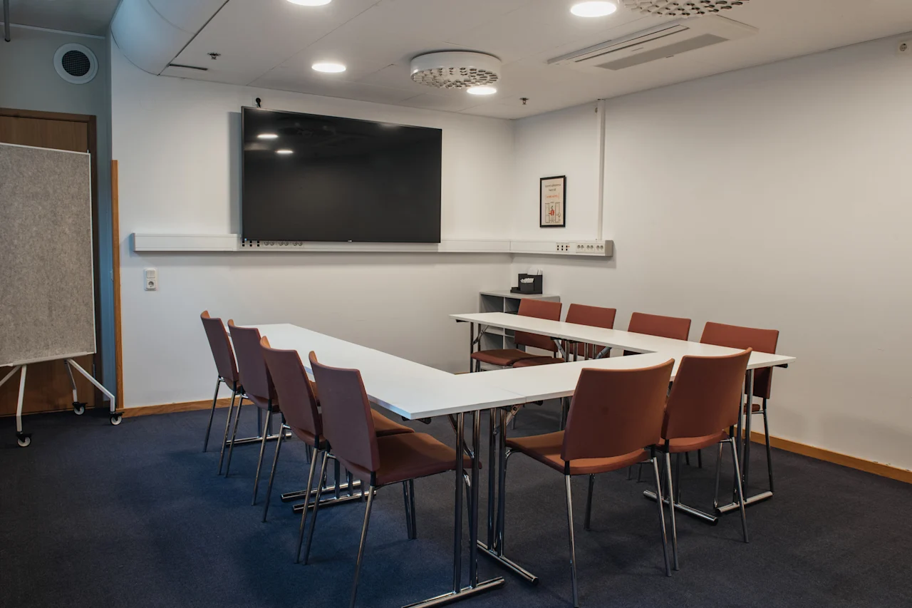 A modern conference room at Home Hotel Uppsala with a U-shaped table, chairs, a large screen, and a whiteboard, ready for a meeting.