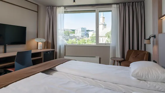 En seng er pænt redt med hvidt sengetøj på et hotelværelse. Et skrivebord med lampe og TV står i nærheden. Et vindue viser udsigt over en by med et kirketårn.