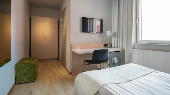En säng vetter mot en väggmonterad TV ovanför ett skrivbord med en stol i ett hotellrum. Ett fönster och en garderob är i närheten, med en grön ottoman längs hallen.