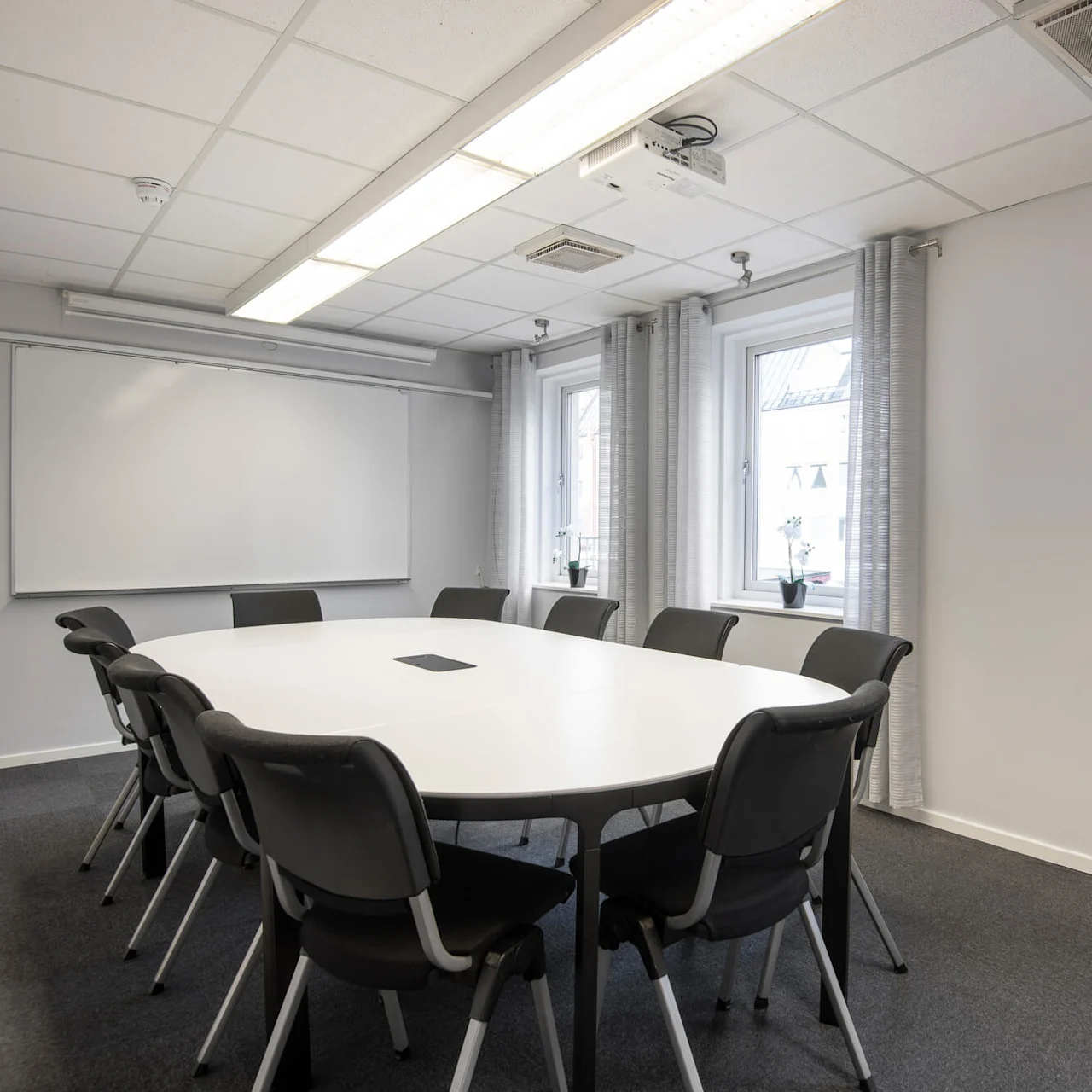 Ett runt bord omgivet av stolar i ett väl upplyst konferensrum, med fönster och en whiteboard på väggarna.