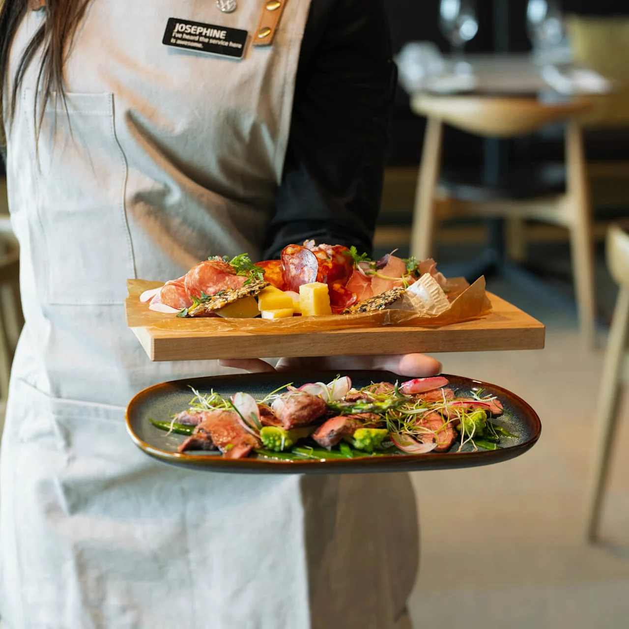 Servitör som bär en träbricka med olika charkuterier och ost, och oval tallrik med skivat kött och grönsaker, i restaurangmiljö. Namnbricka lyder "JOSEPHINE. Jag är ny här, var snäll."