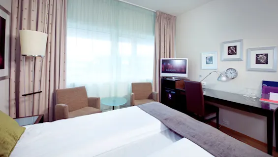 A neatly arranged hotel room with a bed, a television, and various pieces of furniture against a bright, windowed background.