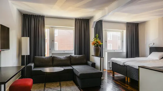 En grå sektionsoffa står i ett modernt hotellrum, intill en säng, med en vas med orange och gröna blommor. Stora fönster med mörka gardiner ger naturligt ljus.