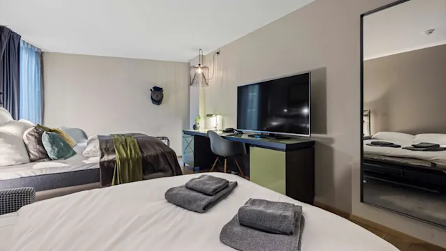 En nøye oppredd seng med foldede håndklær står i et moderne, minimalistisk soverom. En stor TV står på en skrivebord, omgitt av en stol og en lampe. Et speil reflekterer scenen.
