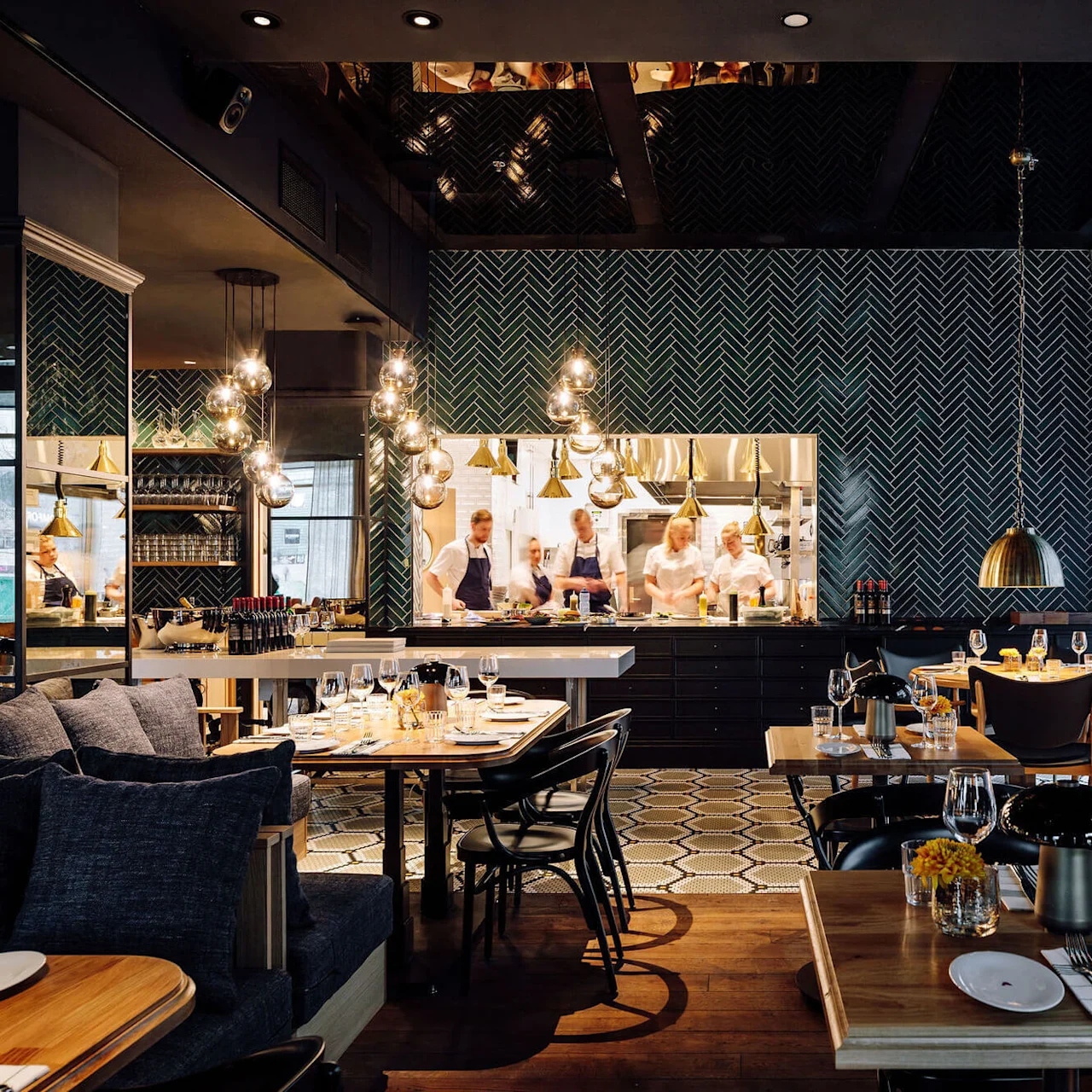 A restaurant features a visible kitchen where chefs prepare food, with modern tables and chairs set for dining in a warmly lit, stylish interior.