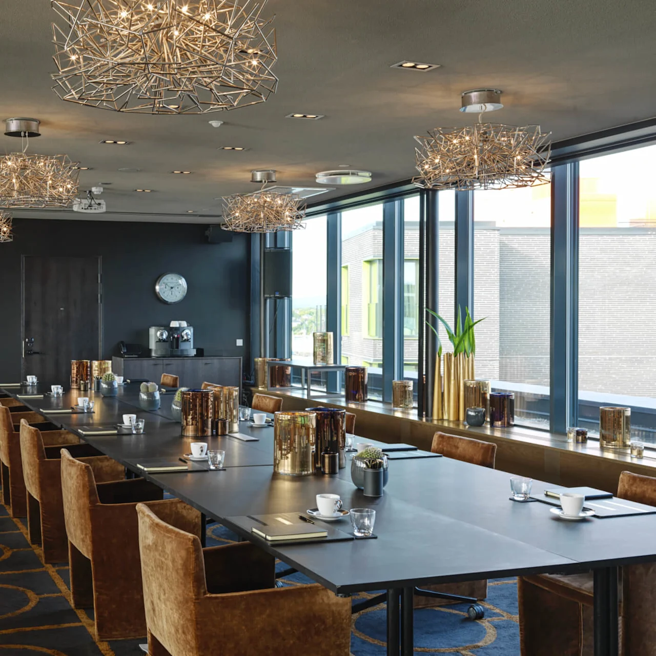 Langt konferansebord med kopper og notatblokker; dekorative lys henger over. Glassvinduer viser en bygning utenfor. Moderne kunst og klokke på veggene; innendørs planter langs vinduskarmen.