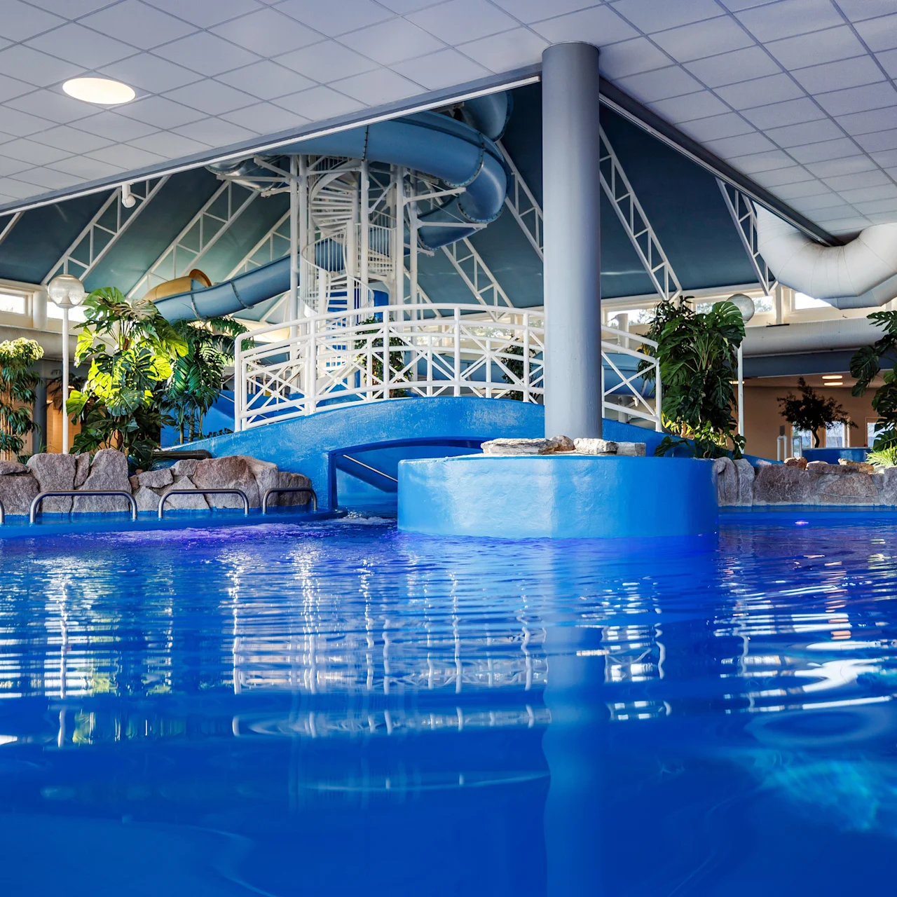 Pool with blue water, surrounded by rocks and plants, features a spiral slide leading into it. Overhead, the ceiling has white tiles and recessed lighting. Lounge chairs are visible nearby.