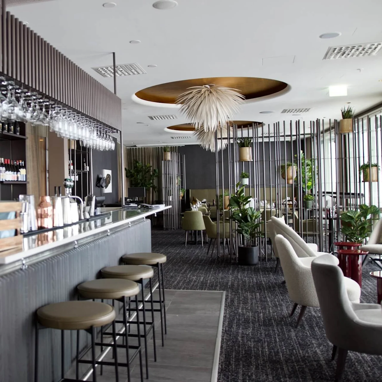 Bar with stools aligned, displaying hanging glasses and various bottles, situated in a modern, stylish lounge with chairs and greenery enhancing the contemporary decor.