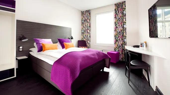 A bed with vibrant purple and orange pillows is in a modern room with dark wood flooring. Floral curtains frame a window beside a sleek desk with a chair.