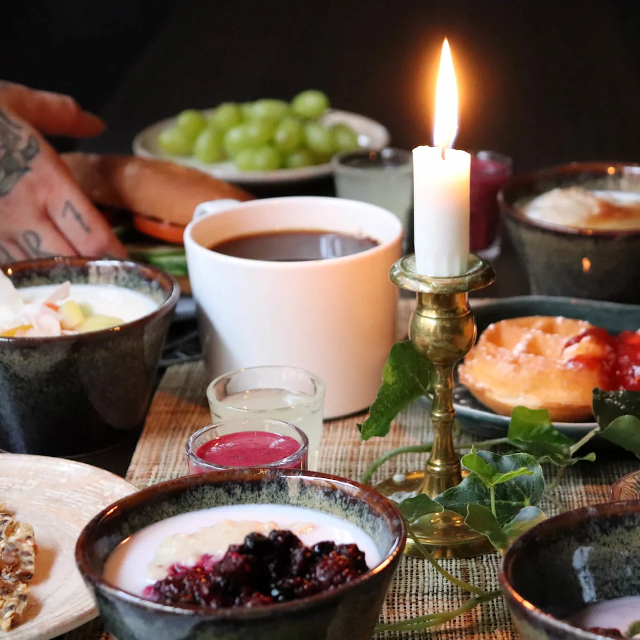 Frokostbord med forskjellige retter, noen holder et glass appelsinjuice, tent lys, koselig atmosfære.