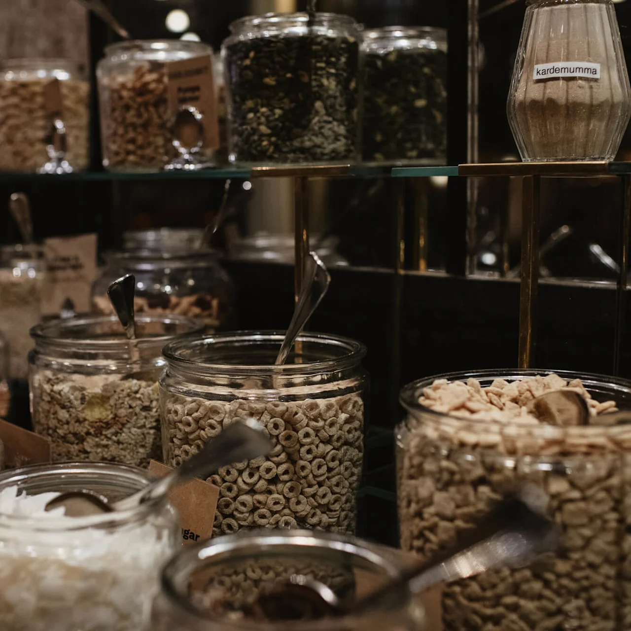Jars filled with various cereals and seeds are arranged on shelves, each with a spoon, in a buffet setting. Visible labels include "kardemumma."