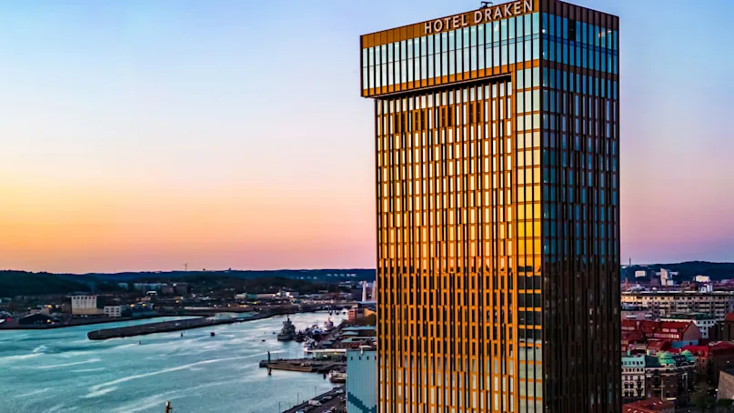 A tall glass building labeled "Hotel Draken" reflects a sunset sky over a waterfront cityscape. Nearby, a river flows alongside urban buildings and docks.