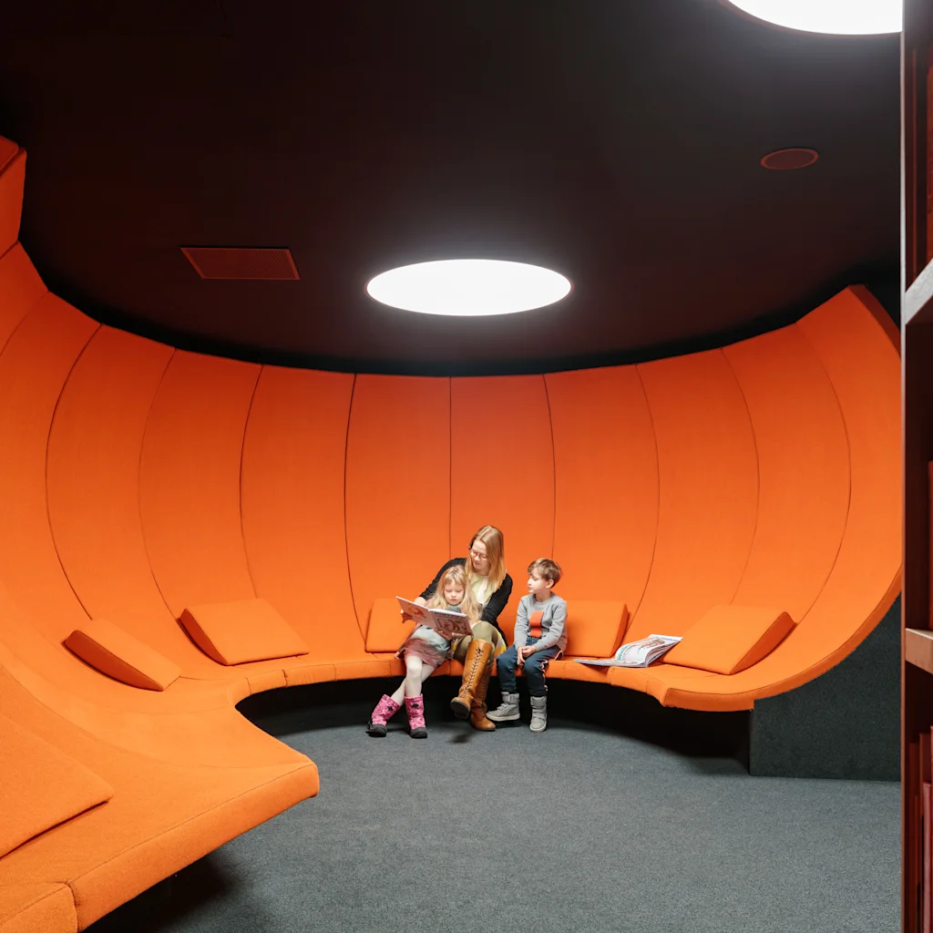 En person leser for to barn i et levende oransje, buet sitteområde på Helsingfors sentralbibliotek Oodi, omgitt av bøker.