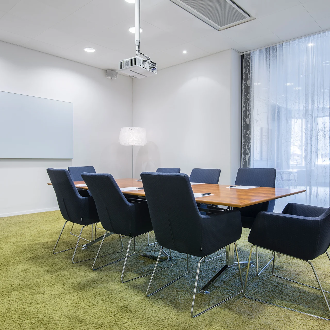 Et rektangulært konferansebord med svarte stoler er arrangert i et møterom. En whiteboard, projektor og gulvlampe kompletterer det moderne, lysbelyste rommet med grønt teppe.