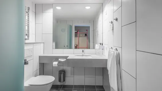 A modern bathroom with a white toilet, a sink with a towel, and a mirror reflecting the opposite wall. Grey tiles cover the floor and walls. Bright overhead lights.