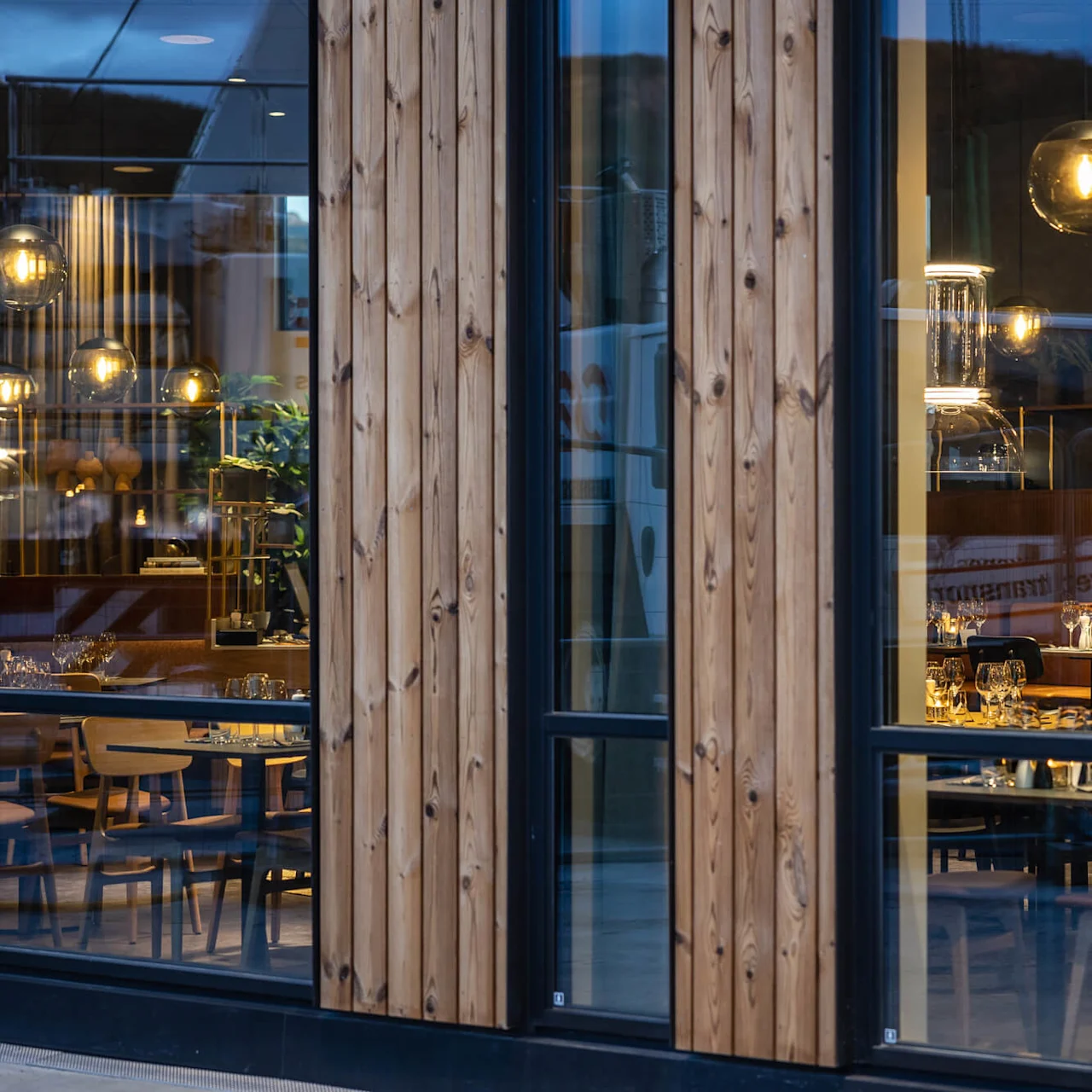 En restaurant synlig gennem store vinduer, med moderne møbler og varm belysning. Træpaneler indrammer scenen. Bordene er dækket med glas og lys i en stilfuld atmosfære.