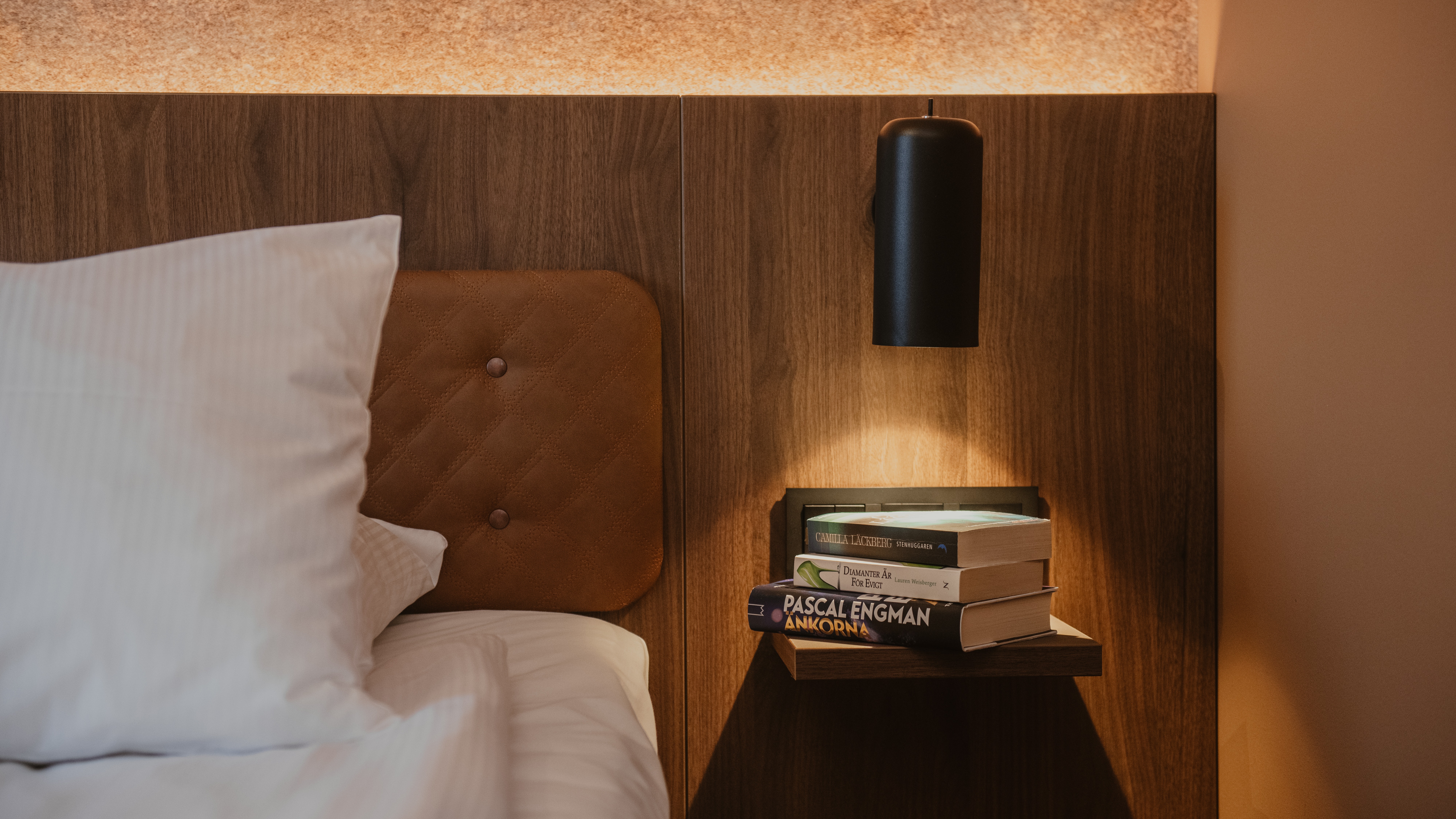 En hyggelig sengeplads med bøger inklusive Pascal Engman Ankorna på en hylde, oplyst af en væglampe, ved siden af en seng.