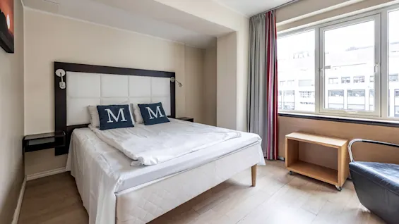 En säng med kuddar som har bokstaven "M" står under ett stort fönster med utsikt över staden. Ett litet träbord och en fåtölj finns i närheten.