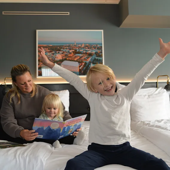 Ett barn med uppsträckta armar sitter glatt på en hotellsäng bredvid en kvinna som läser för ett annat barn. En stadsbild hänger på den mörka väggen i bakgrunden.