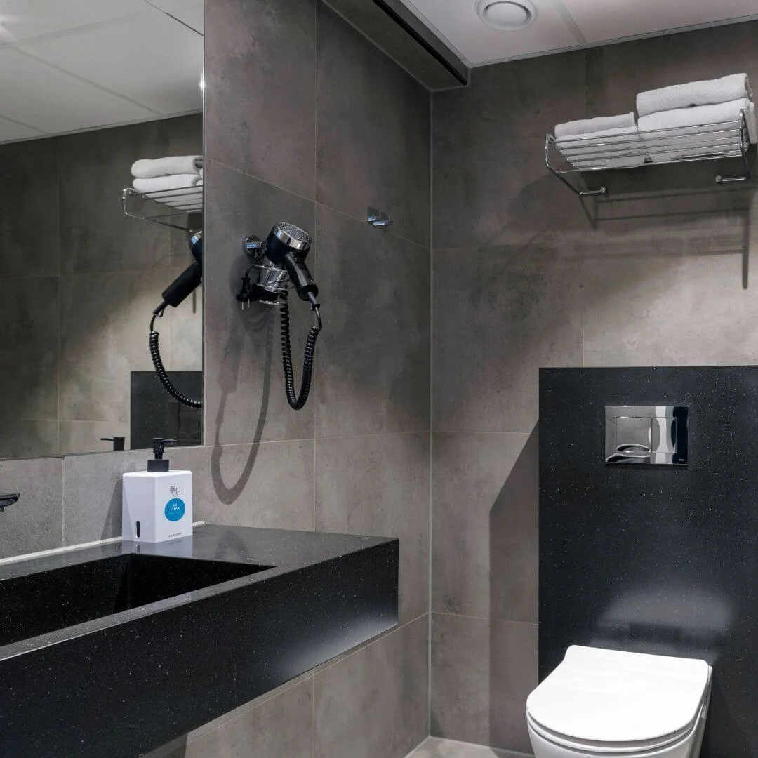 Badeværelse med en moderne vask og vandhane, som spejles i et spejl, en vægmonteret hårtørrer, toilet med metaltryk, og hylde med foldede håndklæder. Grå flisebelagte vægge skaber en smart stemning.