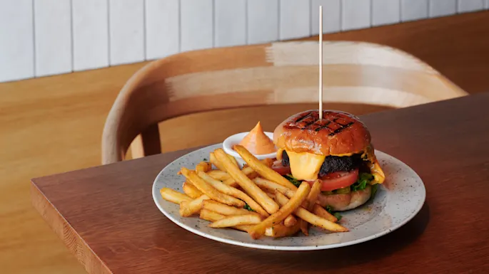 A cheeseburger with lettuce, tomato, and beef patty, served on a plate alongside golden French fries and sauce, sits on a wooden table in a restaurant setting.