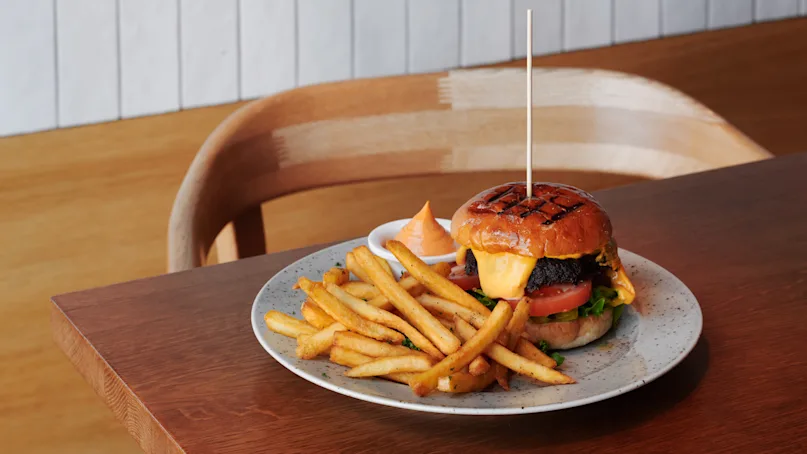 A cheeseburger with lettuce, tomato, and beef patty, served on a plate alongside golden French fries and sauce, sits on a wooden table in a restaurant setting.