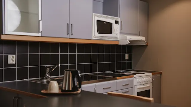 Ett modernt kök med skåp och apparater, inklusive en mikrovågsugn och spis, mot en mörk kaklad backsplash. En vattenkokare och kopp står på bänken i en minimalistisk miljö.