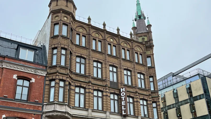 A multi-story brick hotel building with a green spire, featuring a "HOTELL" sign, stands beside a street with people walking and shops, including "Mulberry," visible below.