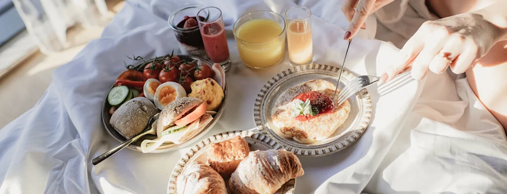 En person i morgenkåpe nyter en deilig frokost på sengen med et utvalg av mat og drikke, badet i naturlig lys.
