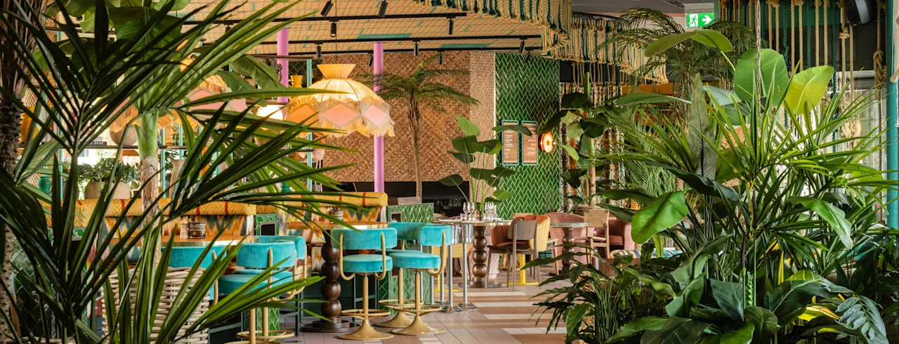 Frodig vegetation fylder en restaurant med farverige siddepladser og hængende stofdekorationer. Grønne fliser, livlige møbler og tropiske planter skaber en livlig, indbydende atmosfære.