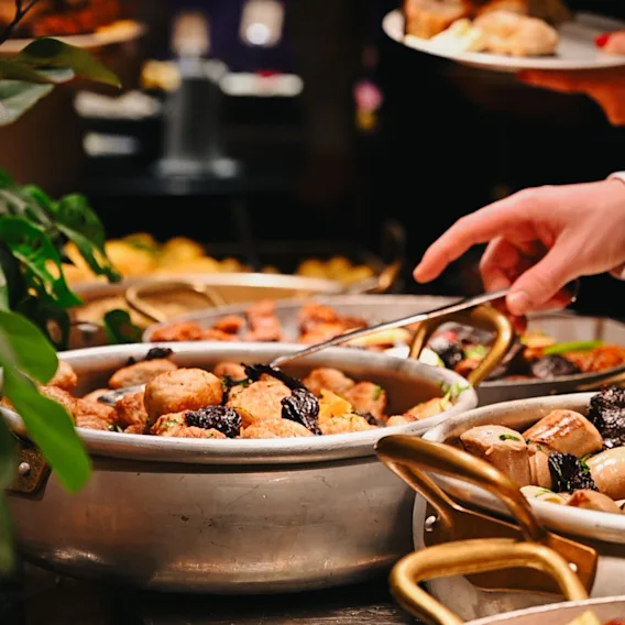 En person forsyner seg med mat fra en buffet, med ulike retter i store gryter.