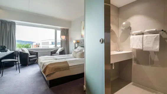 A hotel room with a neatly made double bed, visible through a partially open frosted glass door to an adjacent bathroom. The room has large windows and two gray chairs by a small table.