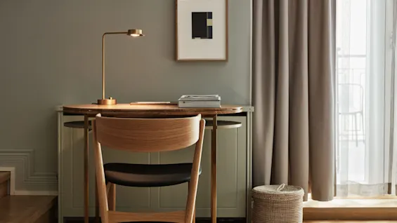 Et skrivebord i tre har en lampe og stabler med bøker, plassert i et minimalistisk rom med lyse vegger og et innrammet abstrakt trykk. Gjennomsiktige gardiner dekker et vindu.