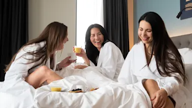 Tre kvinner i hvite badekåper ler og nyter drinker på en seng i et lyst hotellrom, med frokost anrettet i nærheten.