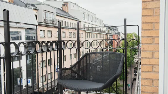 A black chair sits on a balcony overlooking a city street, surrounded by iron railings with circular patterns, with buildings and trees in the background.