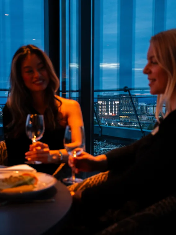 To personer skåler med vinglass ved et restaurantbord foran store vinduer om kvelden med bylys i bakgrunnen.